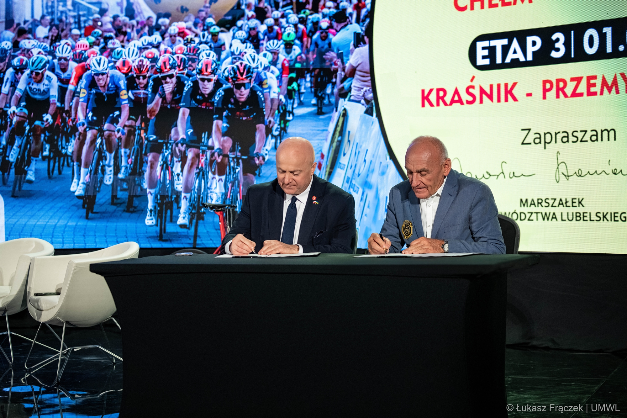 Na zdjęciu widzimy Marszałka Województwa Lubelskiego Jarosława Stawiarskiego i Czesława Langa dyrektora generalnego wyścigu Tour de Pologne, którzy podpisują umowę na organizację tego wyścigu. Za ich plecami, na dużym ekranie zdjęcie przedstawiające grupę kolarzy oraz trasa jednego z etapów.