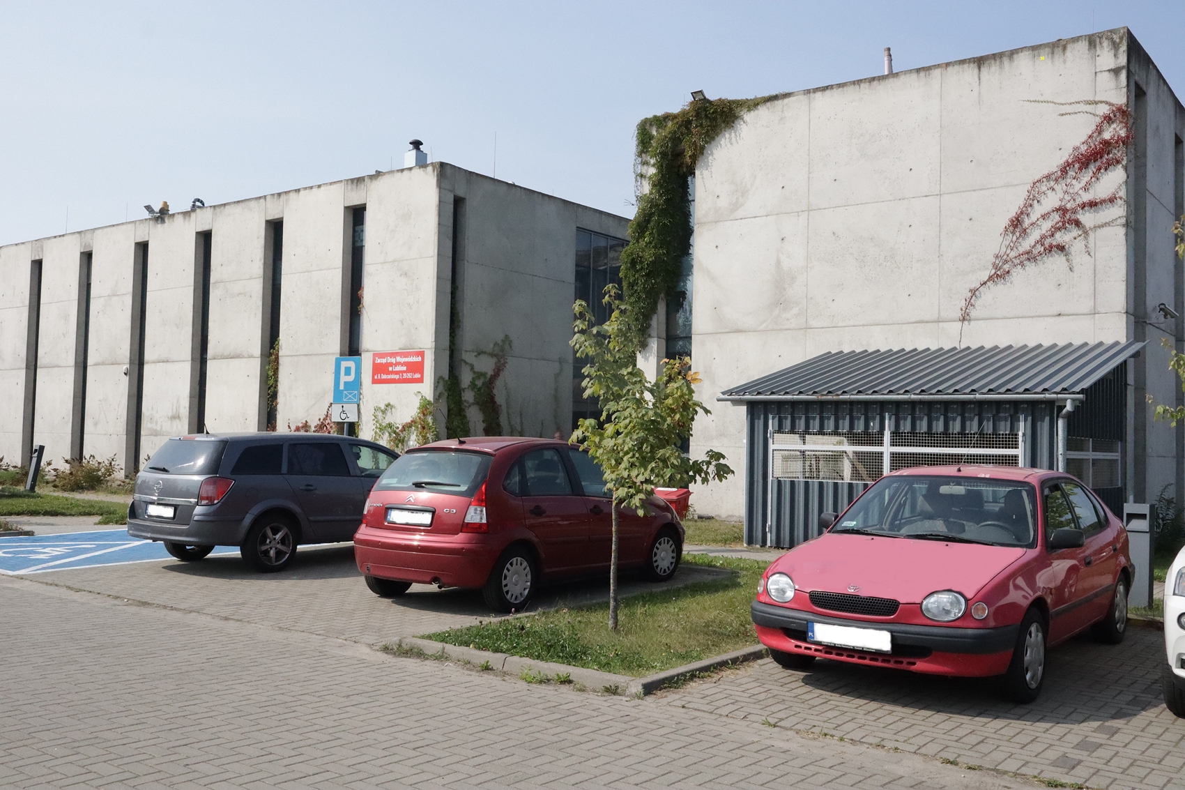 Zdjęcie przedstawia budynek Lubelskiego Parku Naukowo Technologicznego, w którym swoją siedzibę ma Zarząd Dróg Wojewódzkich w Lublinie. Budynek wykonany jest w całości z szarego betonu. Z każdej strony znajdują się podłużne, wąskie okna. Przed budynkiem widzimy ustawione na parkingu samochody osobowe.