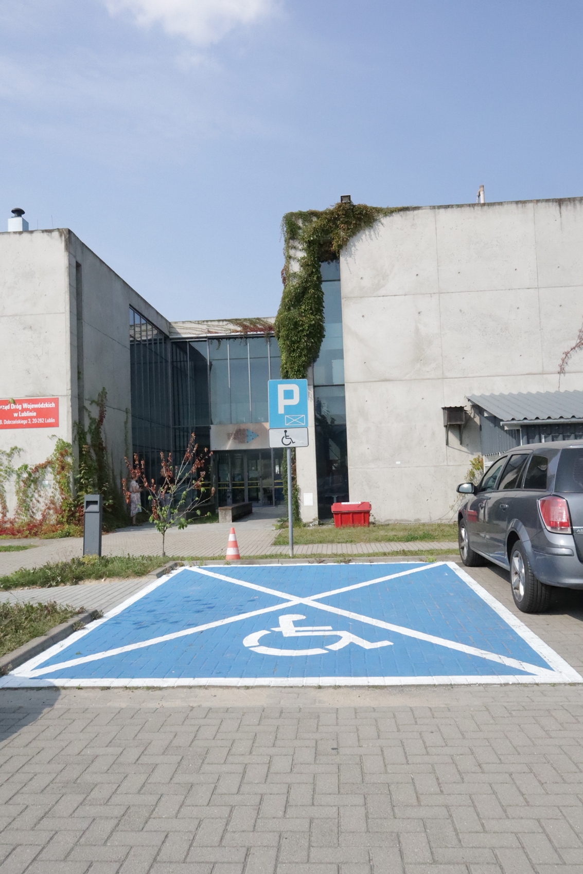 Na zdjęciu widzimy miejsce parkingowe dostosowane dla osób ze specjalnymi potrzebami. Miejsce to znajduje się około 12 metrów od wejścia głównego do budynku. Miejsce parkingowe oznaczone jest kolorem niebieskim oraz namalowanym białym znakiem X a także oznaczeniem osoby poruszającej się na wózku.