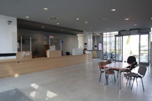 Na zdjęciu widzimy główny hall w Zarządzie Dróg Wojewódzkich w Lublinie, który znajduje się na tym samym poziomie co parking. Hall ma około 32 metrów kwadratowych. Podłoga na nim jest z płytek w kolorze szarym. Po lewej stronie widzimy biuro podawcze Zarządu Dróg Wojewódzkich w Lublinie oraz miejsce w którym przebywa ochrona. Blat w biurze podawczym znajduje się na wysokości, która umożliwia podjechanie osobie na wózku i wykonany jest z jasnego drewna.