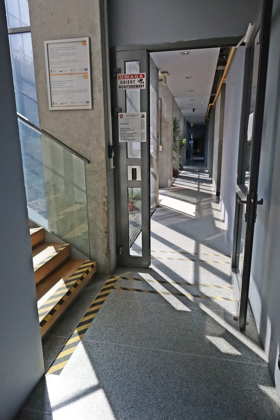 Na zdjęciu widzimy na dojście do schodów w budynku Zarządu Dróg Wojewódzkich w Lublinie oraz przejście z głównego hallu na korytarz prowadzący do pokoi pracowniczych. Na podłodze żółto czarną taśmą wyklejone są pasy zarówno przed wejściem na schody jak i przed drzwiami wejściowymi na korytarz.