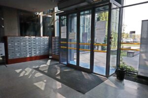 Na zdjęciu widzimy drzwi wejściowe do budynku Zarządu Dróg Wojewódzkich w Lublinie. Są one szklane z jasno szarą ramą. Na wysokości 75 centymetrów i 100 centymetrów wyklejone są dwa pasy w kolorze żółtym w celu kontrastu. Drzwi otwierają się samoczynnie dzięki zamontowanej nad wejściem czujce.